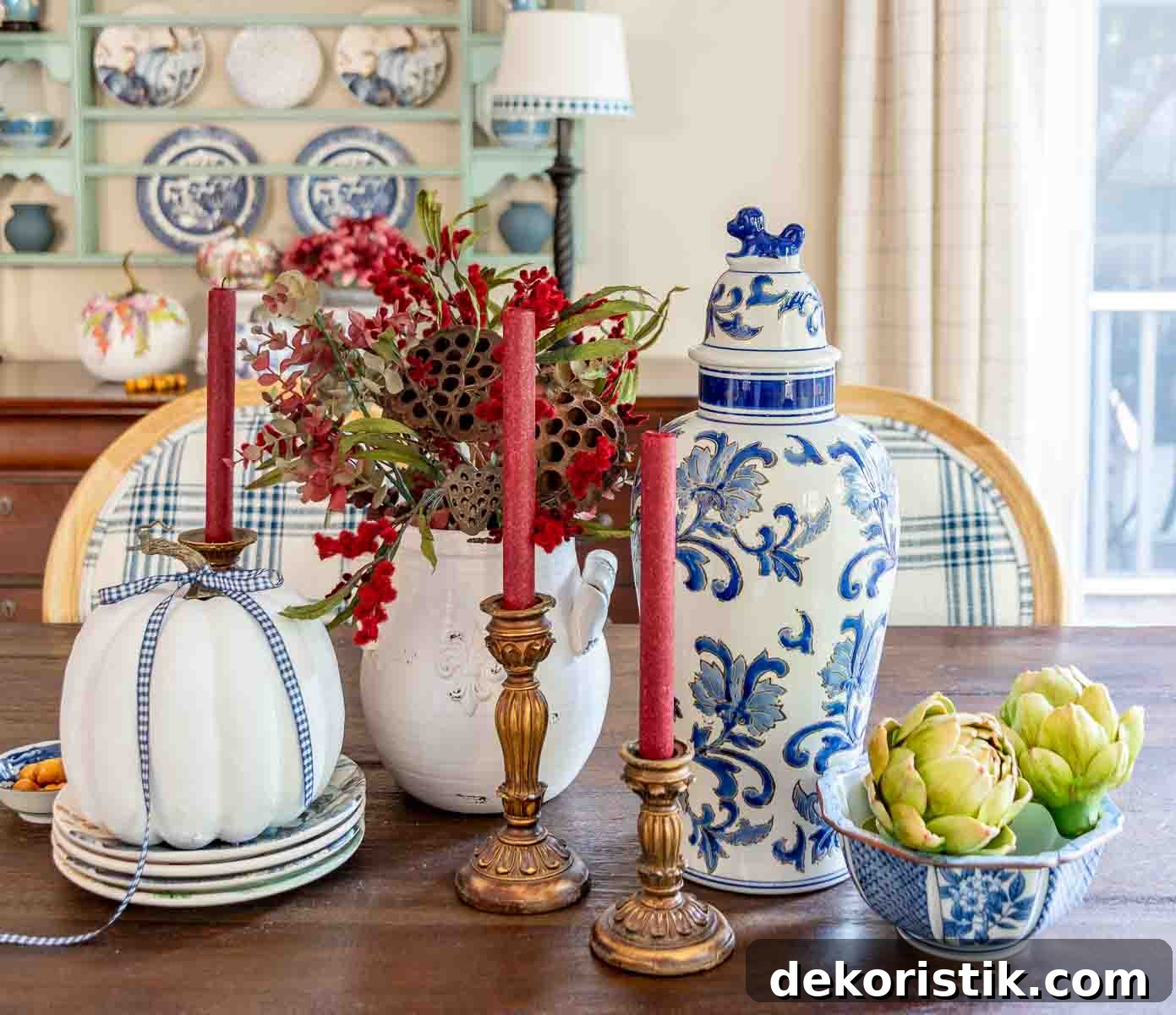 a diy fall table centerpiece using blue, white and burgundy colors, consisting of a large lidded urn, a floral arrangement, and a large white pumpkin on a stack of fall plates