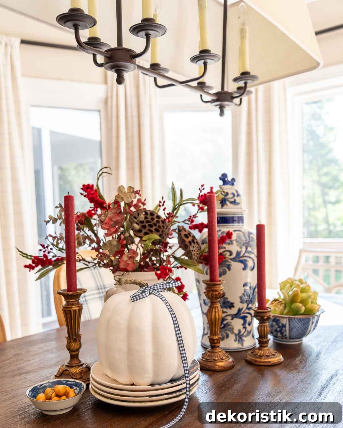 a diy fall table centerpiece using blue, white and burgundy colors, consisting of a large lidded urn, a floral arrangement, and a large white pumpkin on a stack of fall plates