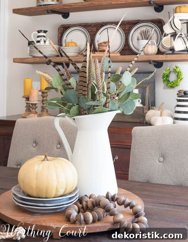 A white pitcher is filled with eucalyptus leaves, feathers and it is on a wooden tray that is round on the table.