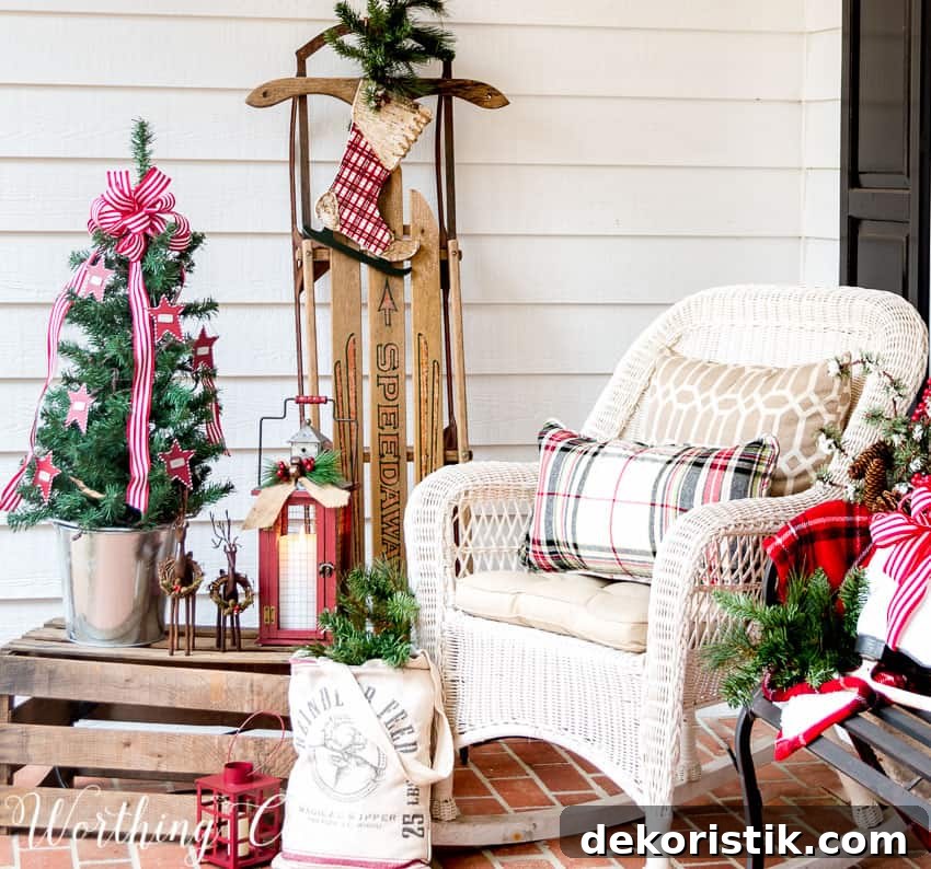 A porch decorated with a mini Christmas tree, a white rocking chair with a buffalo checked pillow and a wooden sled.