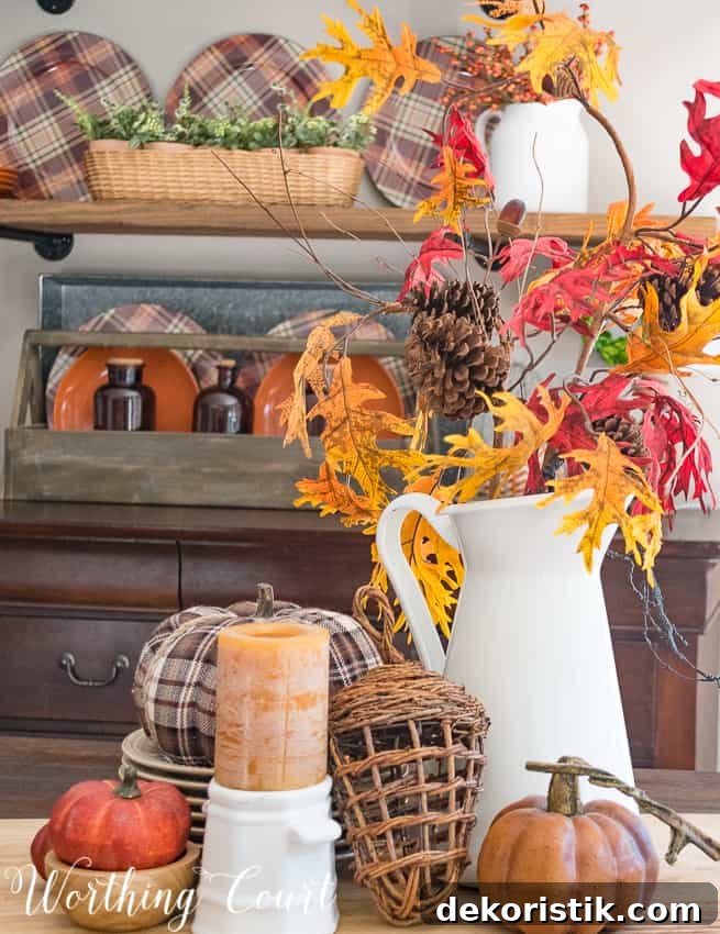 A white pitcher on the table filled with fall leaves, a plaid pumpkin, a candle and small pumpkins.