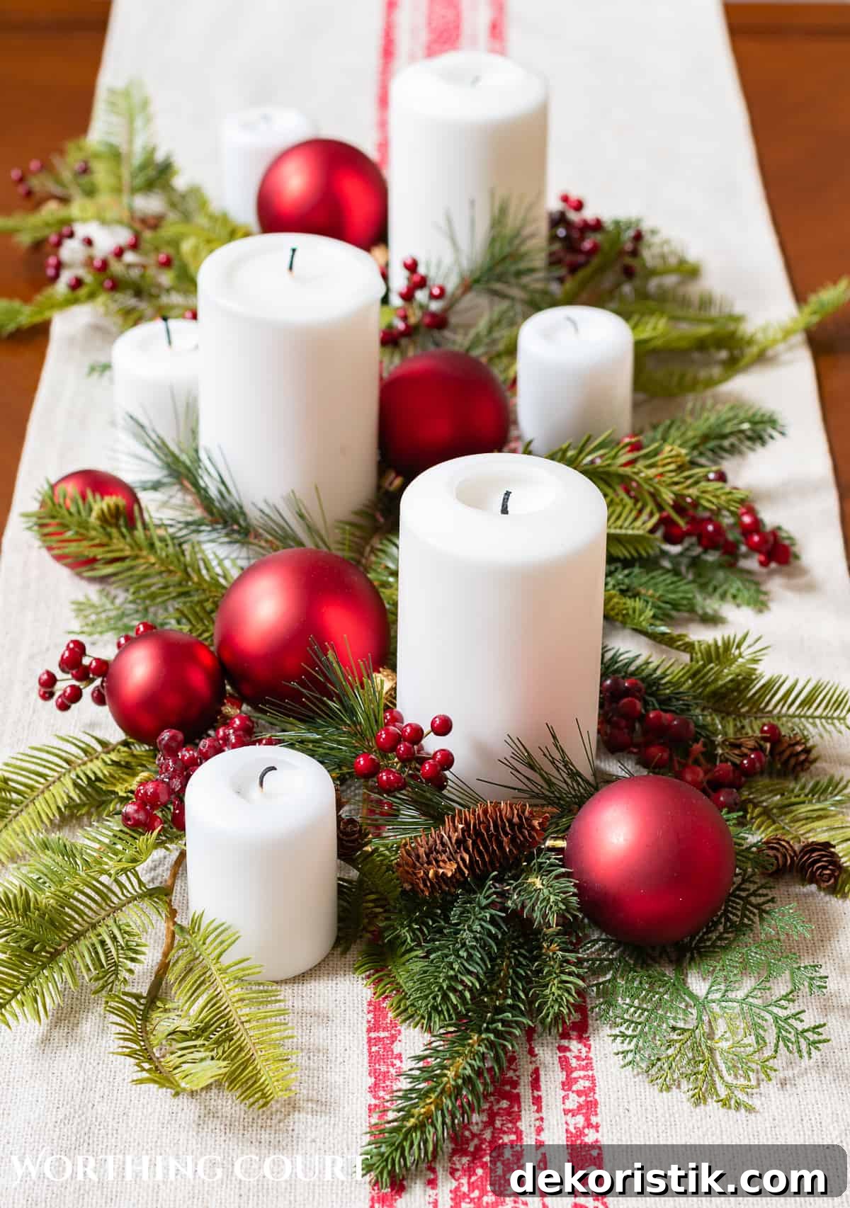 Assorted white candles on a coffee table with faux greenery and red Christmas ornaments on top of a table runner