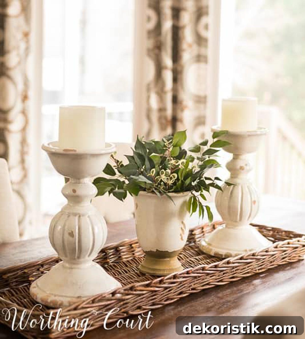 greenery in a white vase between two chunky white candlesticks on a long wicker tray on a dining table