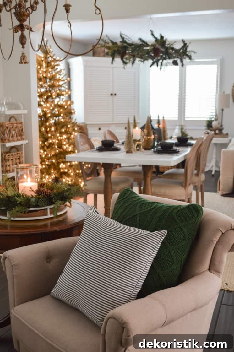 A neutral armchair with a green throw pillow on it. There is a lighted Christmas tree in the dining room.