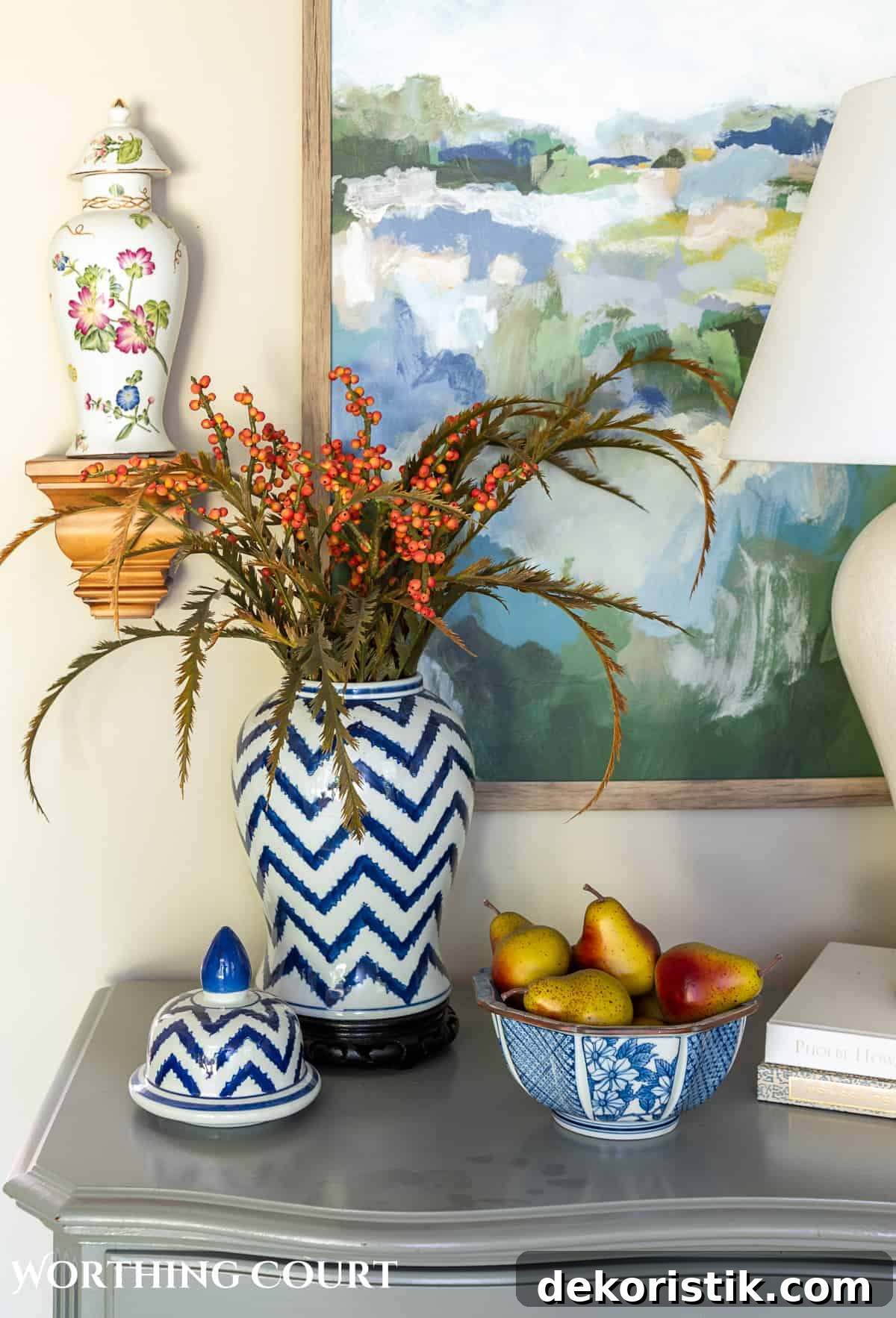 blue and white ginger jar with faux fall stems and a bowl of fake pears beside a white lamp on a gray chest