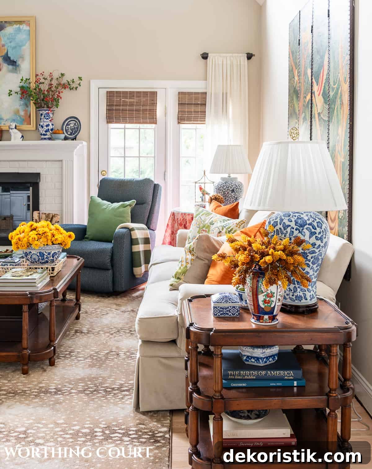side view of beige sofa and blue chair beside white brick fireplace with fall decorations and a blue and white lamp on a brown wood end table