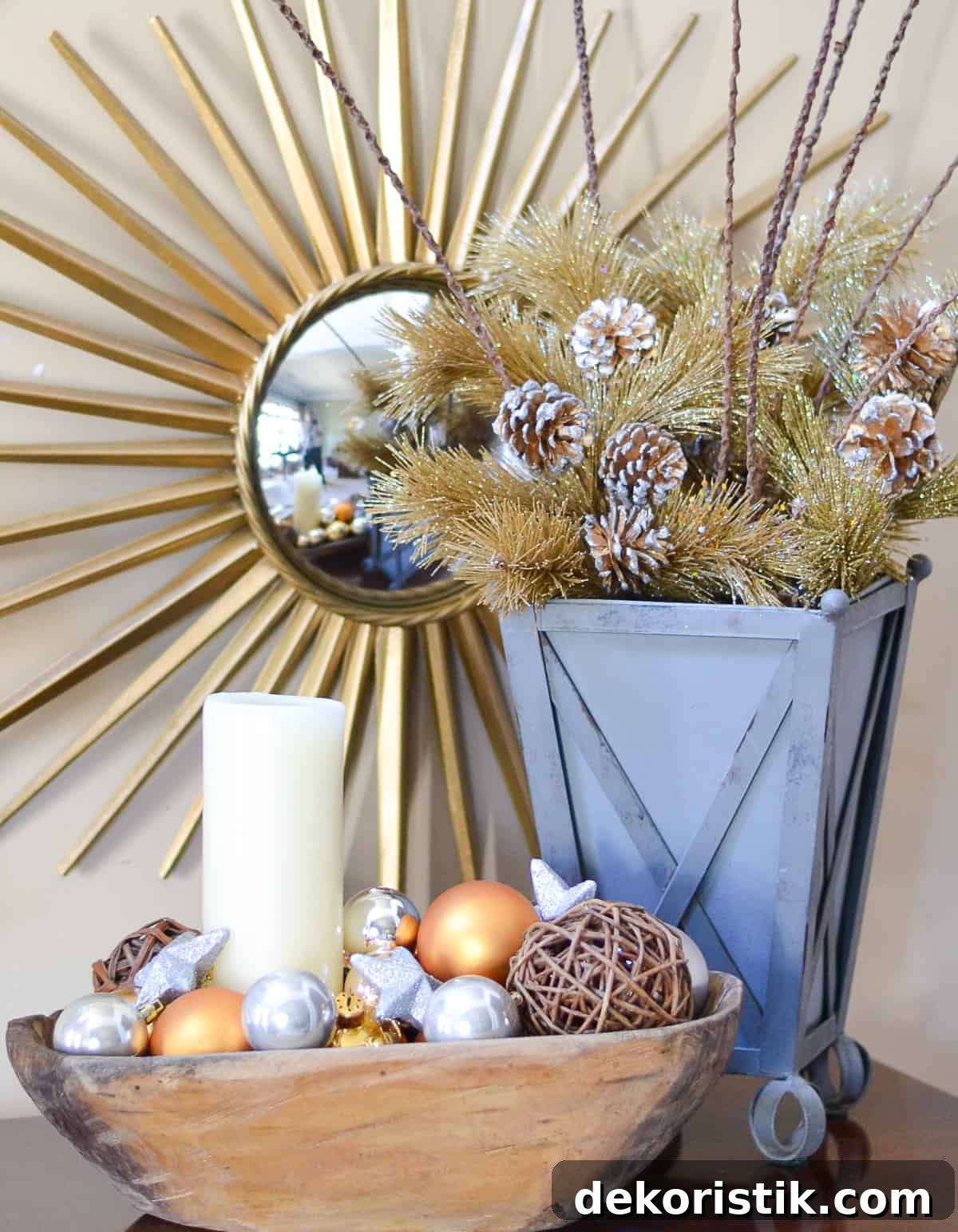 gold and silver ornaments and a wood bowl and a gold and pine cone arrangement in a gray container in front of a sunburst mirror