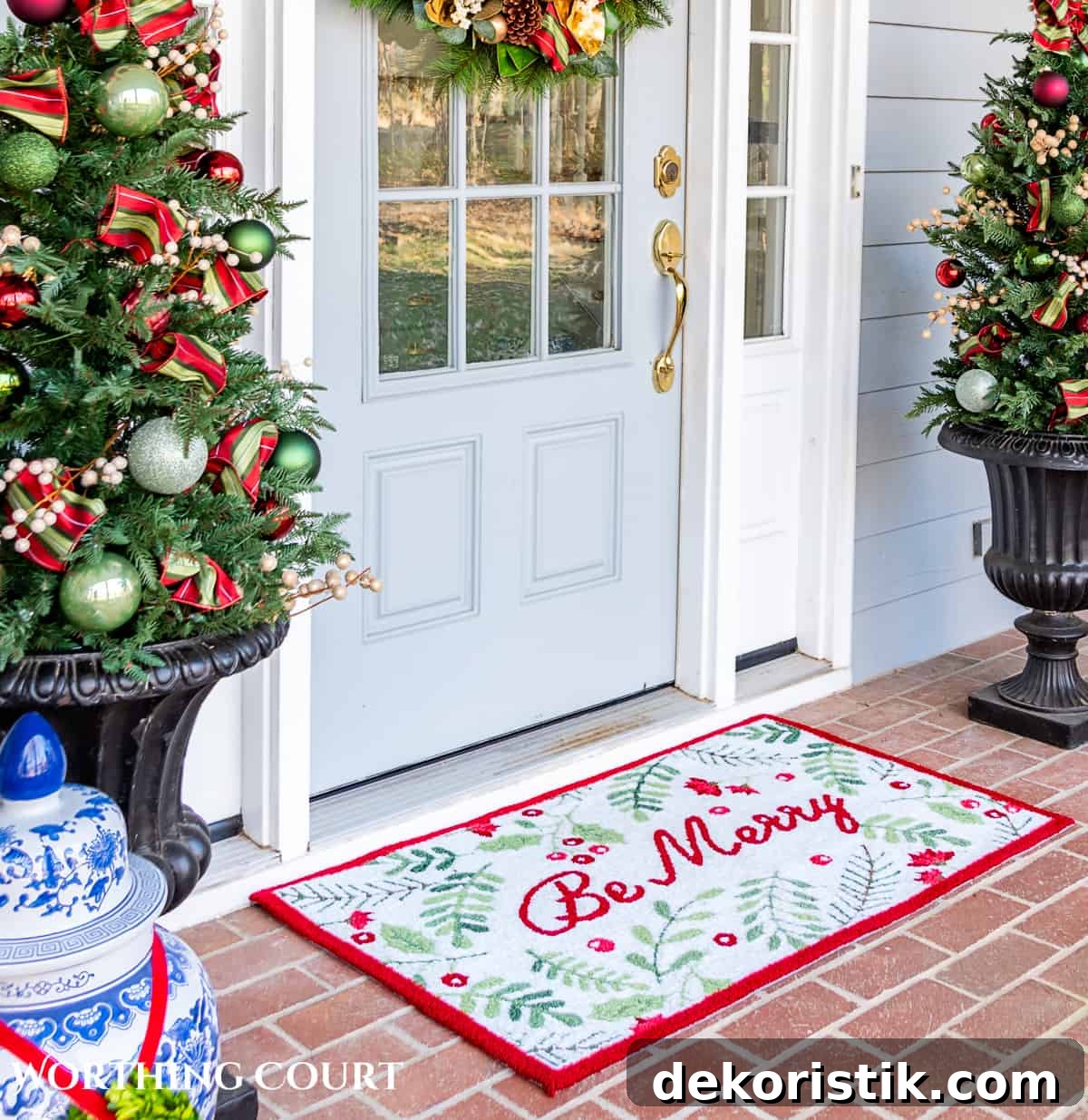 red, green and white Christmas welcome mat with the words "Be Merry" in front of a gray front door flanked by small Christmas trees in urns