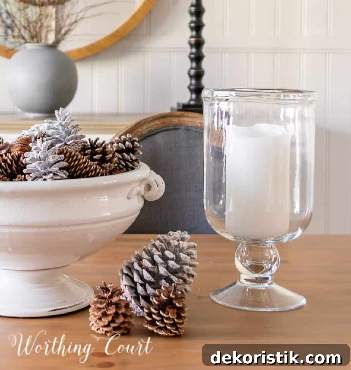large white pedestal bowl on a dining table decorated for winter with pinecones and flanked by glass hurricanes