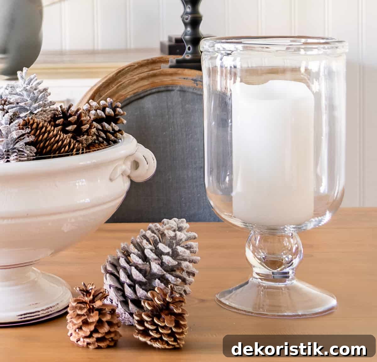 large white pedestal bowl on a dining table decorated for winter with pinecones and flanked by glass hurricanes