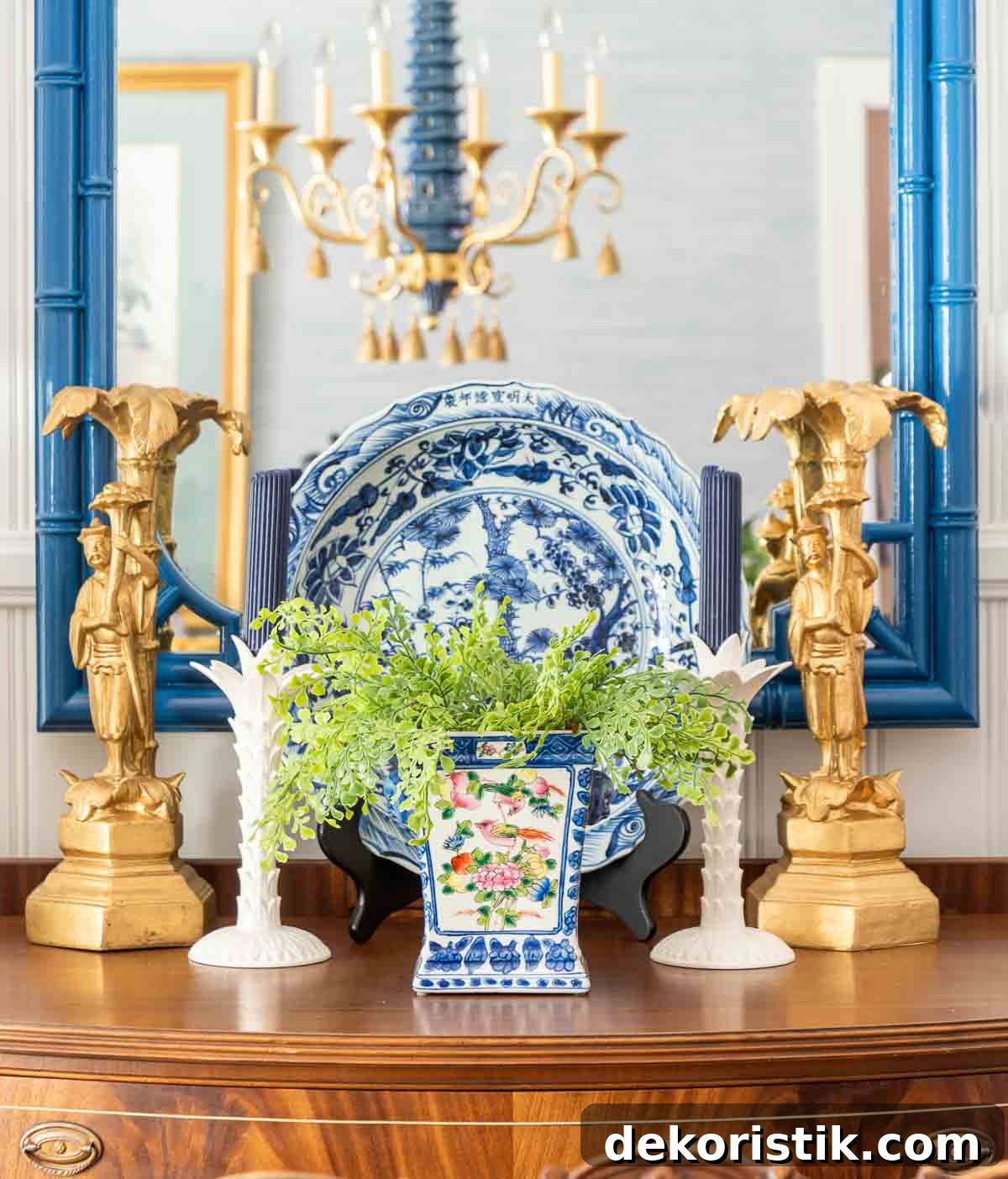 two gold chinese men figurines, two white candlesticks with blue candles, a large round blue and white platter and a chinoiserie planter with faux greenery arranged in a vignette in front of a mirror