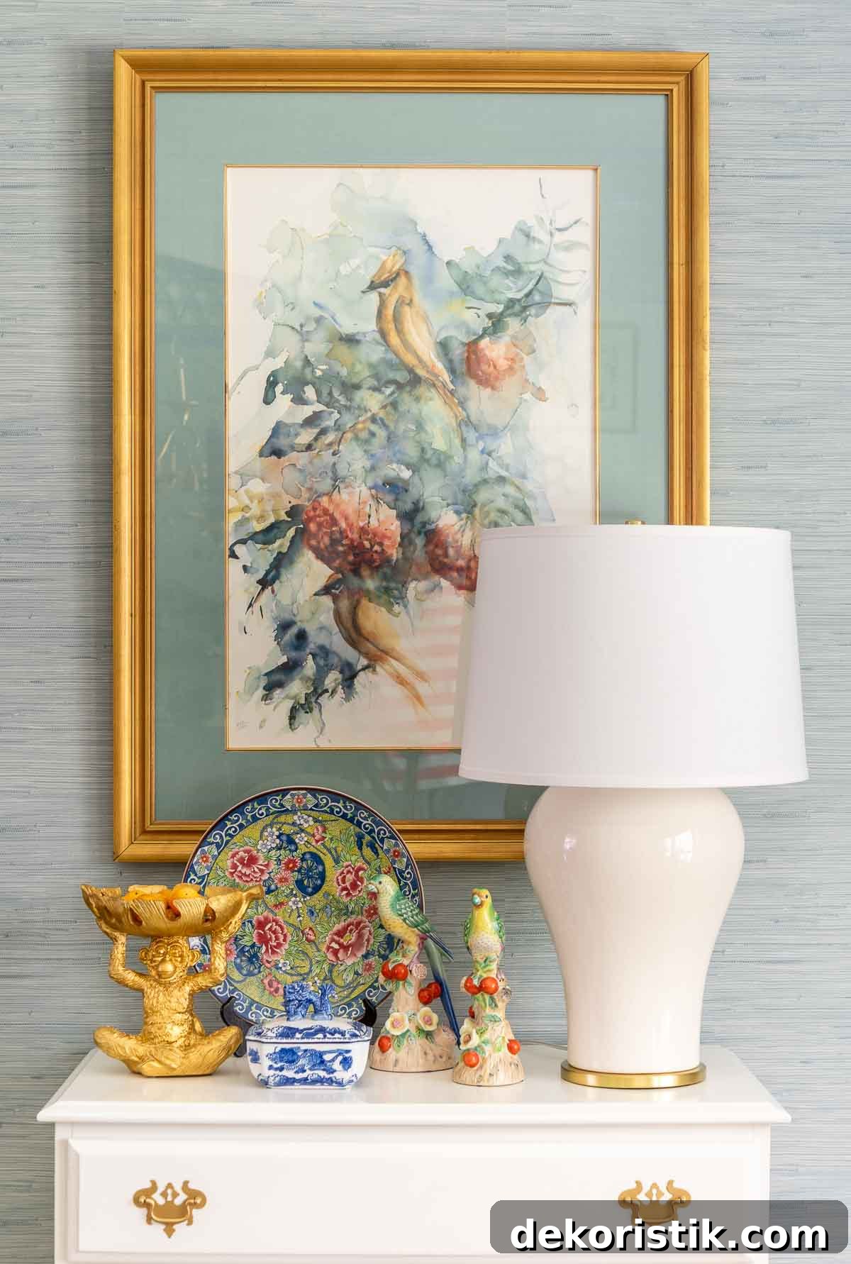 white lamp and chinoiserie accessories on a white chest, underneath traditional style artwork against a wall with blue grasscloth wallpaper