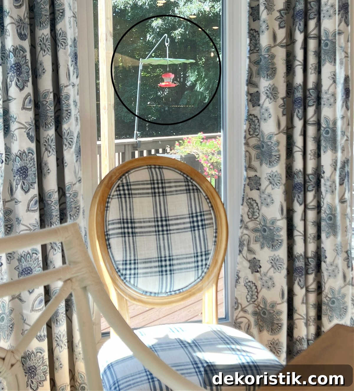 View through a window of a hummingbird feeder with a blue and white chair sitting in front of the window