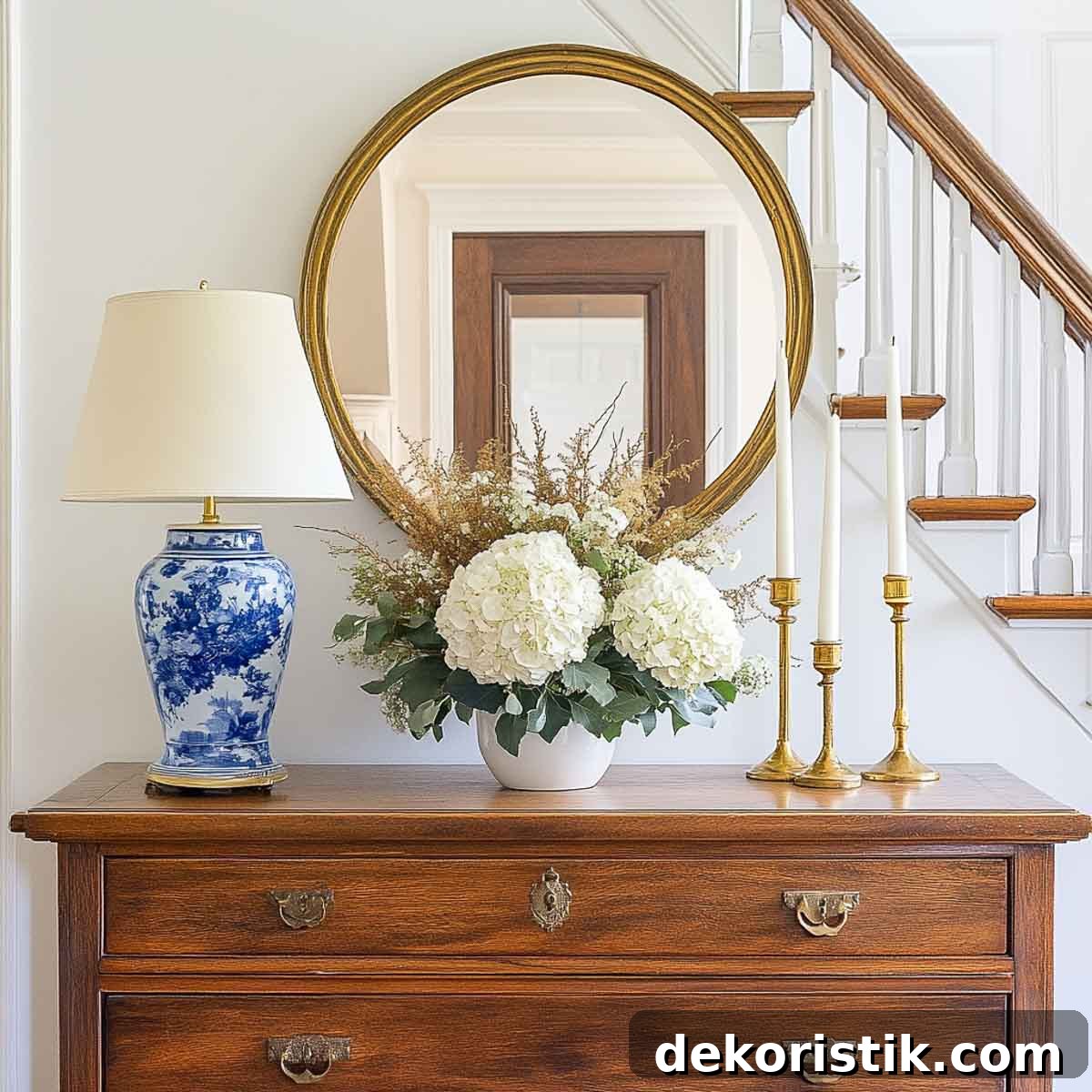 foyer console table with a round mirror above, a blue and white lamp on one end, brass candlesticks on the other and a floral arrangement in the center
