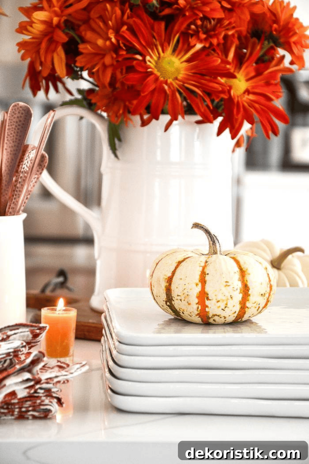 white pitcher filled with fall flowers behind a stack of white dishes with a small pumpkin on top