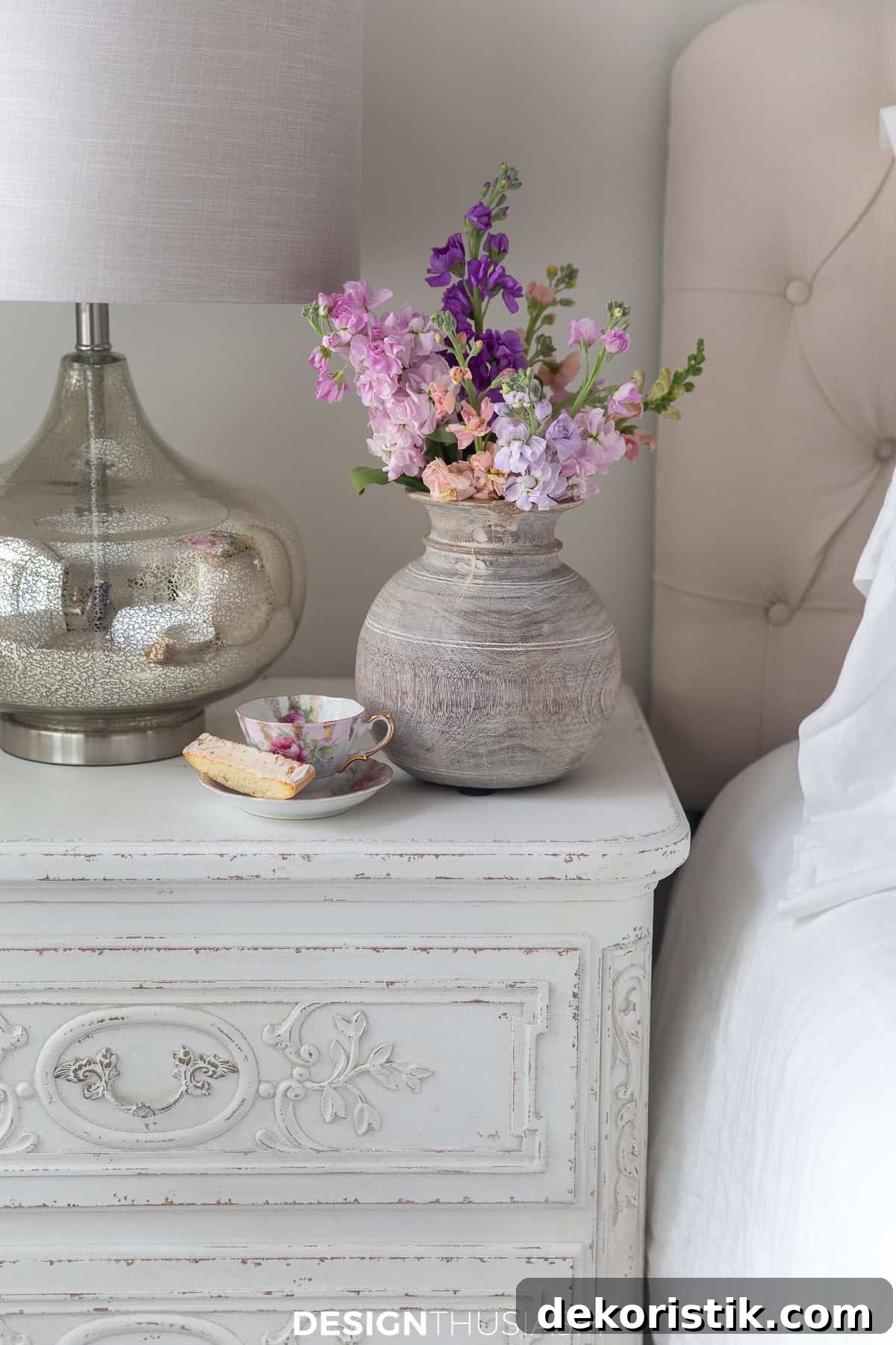 corner of a white nightstand beside a bed with a vase of pink flowers