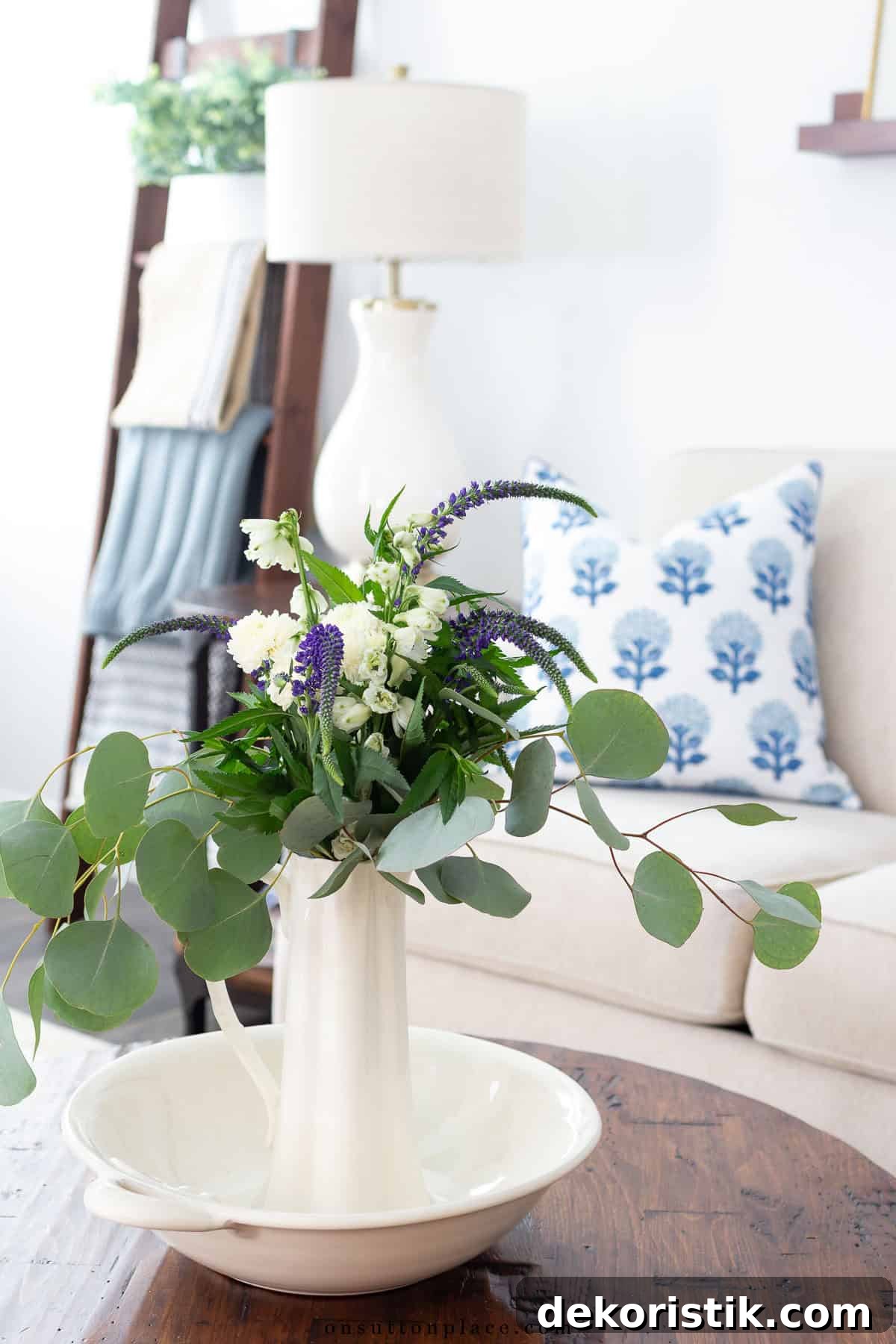 greenery in white vessels on a wood coffee table in front of a white couch with a blue and white throw pillow