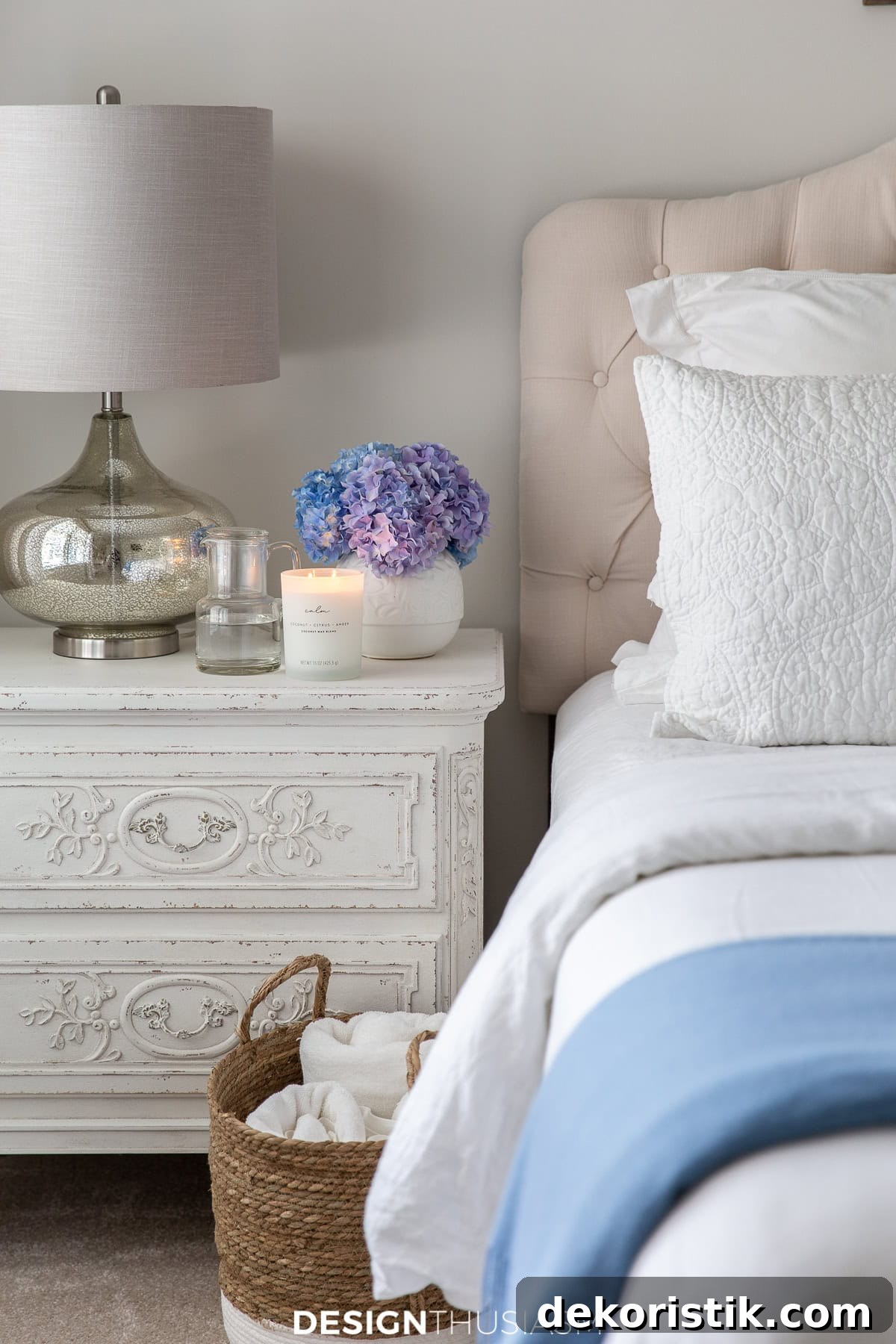 top corner of a bed with white linens and a neutral upholstered headboard beside a white nightstand with a mirrored table lamp