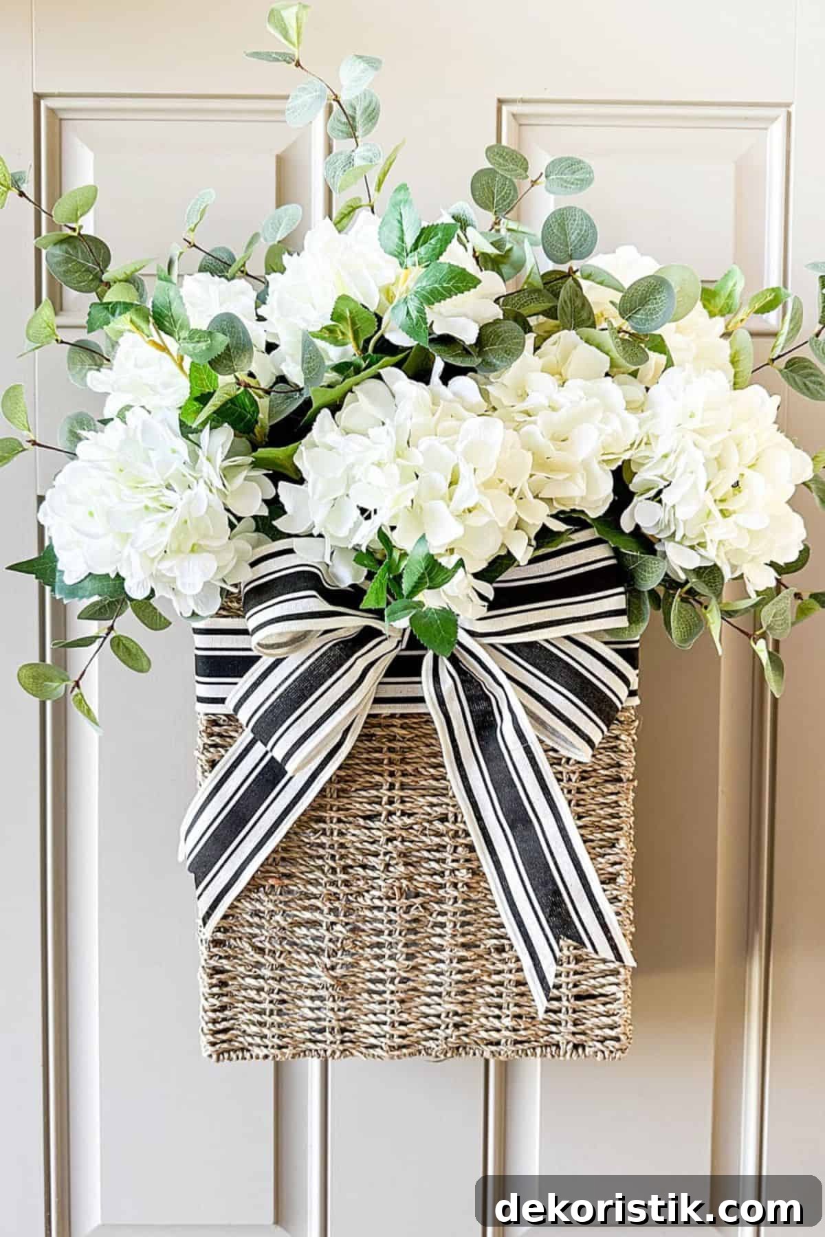 wicker basket hanging on a beige front door filled with faux white geraniums and embellished with a black and white striped bow