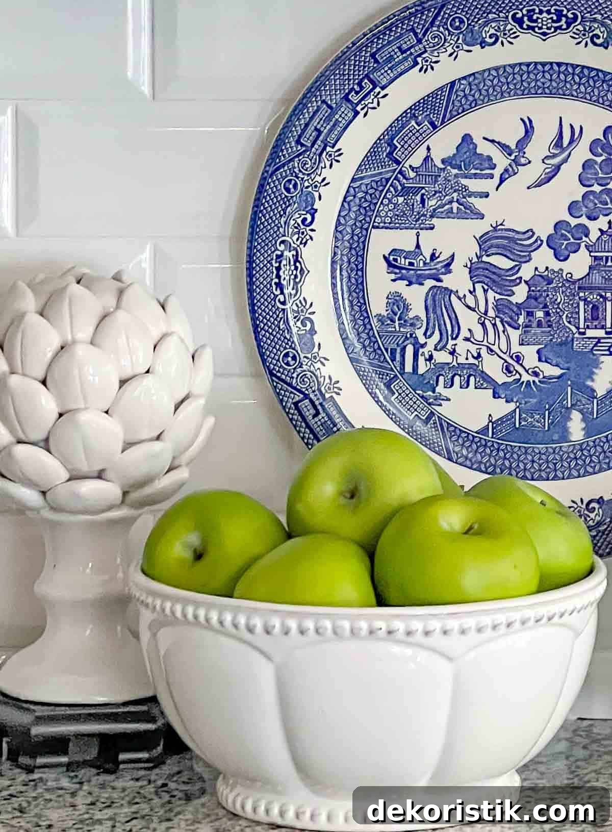 Green apples in a white bowl and white artichoke on a kitchen counter with a blue and white plate
