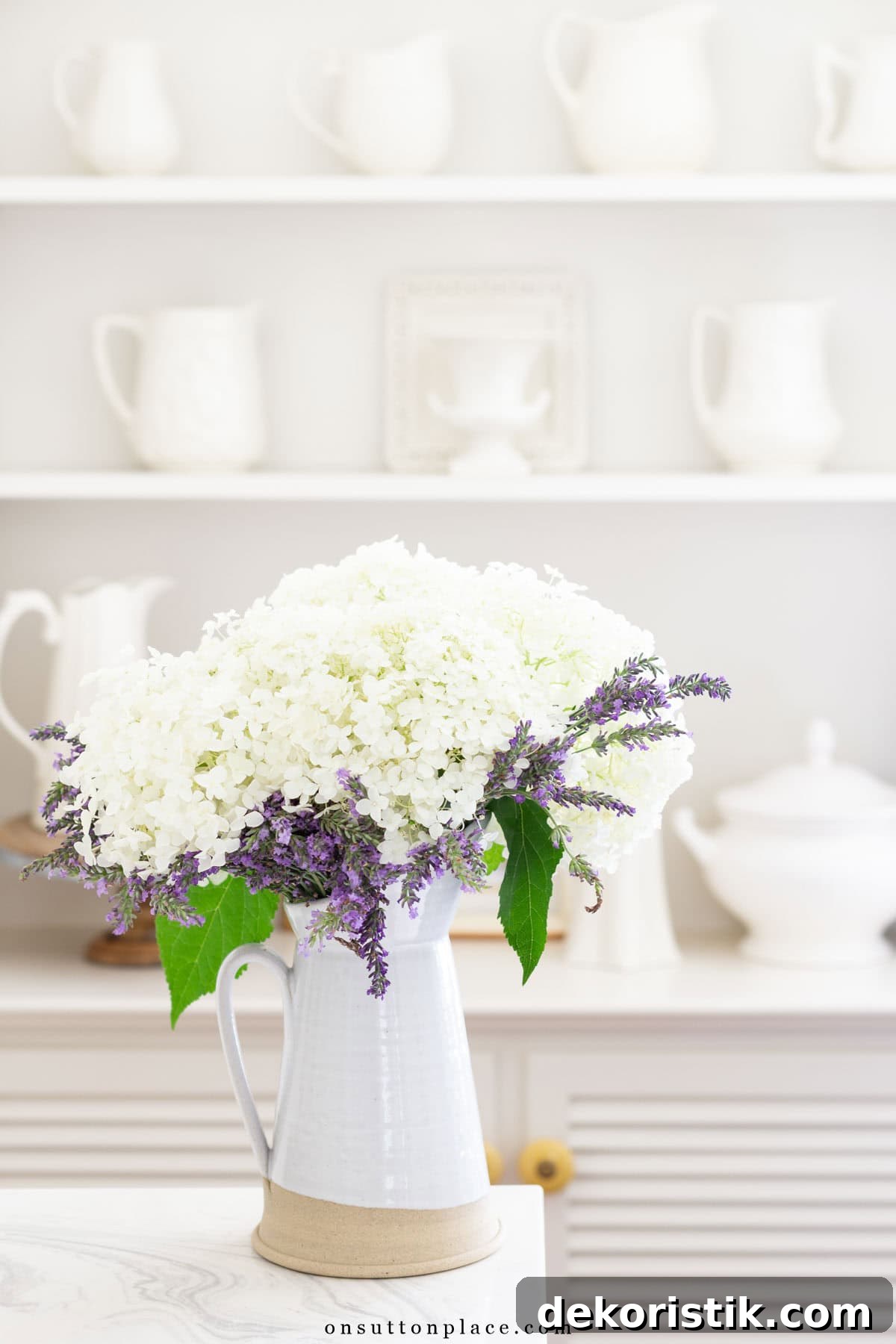white cut flowers in a white vase in front of shelves containing white dishes